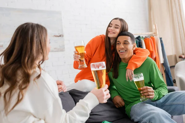 Smiling woman hugging cheerful african american man while holding glass of beer and looking at friend — Photo de stock