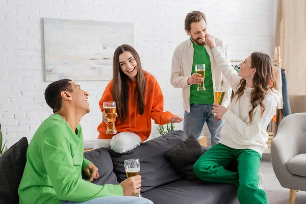 Amigos multiétnicos felizes segurando copos de cerveja enquanto conversa na sala de estar — Fotografia de Stock