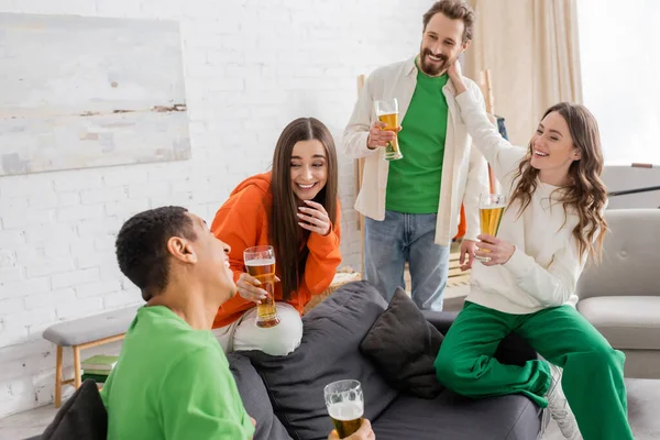 Alegres amigos multiétnicos sosteniendo vasos de cerveza mientras charlan en la sala de estar - foto de stock