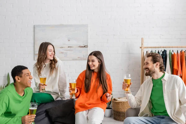 Alegres amigos multiétnicos sosteniendo vasos de cerveza mientras sonríen en el salón - foto de stock