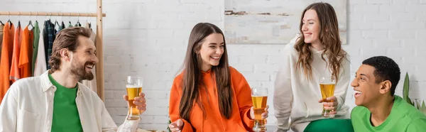 Веселі багатоетнічні друзі тримають келихи пива, посміхаючись у вітальні, банер — стокове фото