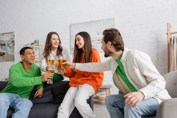 Fröhliche multiethnische Freunde klimpern im Wohnzimmer mit Biergläsern — Stockfoto