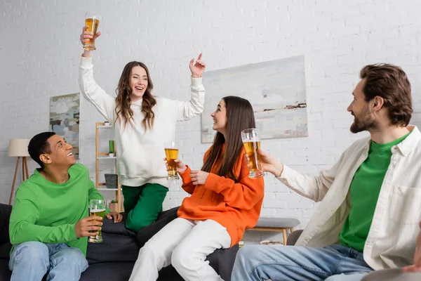 Excited woman with glass of beer gesturing near interracial friends in living room — Stockfoto