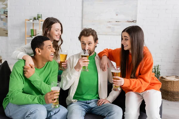 Fröhliche interrassische Gruppe von Menschen, die einen bärtigen Freund beim Bier im Wohnzimmer anschauen — Stockfoto