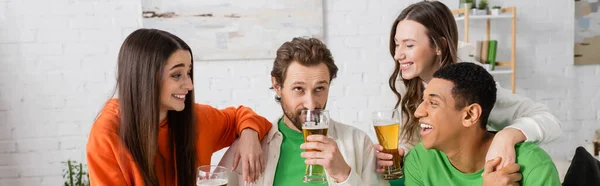 Щаслива міжрасова група людей, які дивляться на бородатого друга, що п'є пиво у вітальні, банер — стокове фото