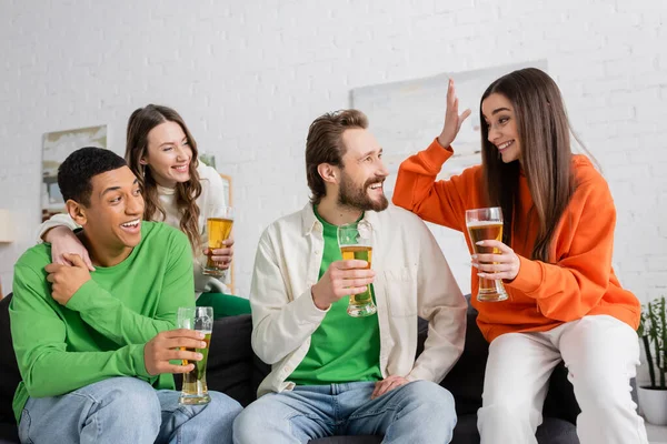 Fröhliche gemischtrassige Menschen schauen fröhliche Frau gestikulierend an, während sie Bier im Wohnzimmer hält — Stockfoto