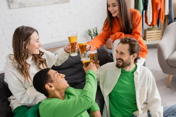 Alegre grupo multicultural de amigos tintineando vasos de cerveza y mirándose en la sala de estar - foto de stock