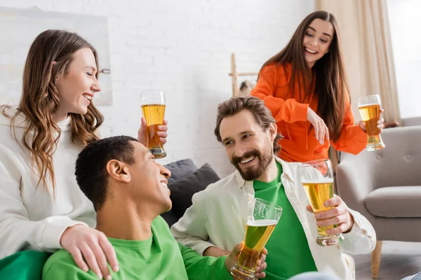 Cheerful multicultural group of friends holding glasses of beer while chatting in living room — Stockfoto