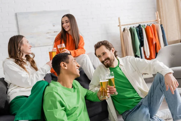 Cheerful multicultural men and women clinking glasses of beer while chatting in living room — Stockfoto