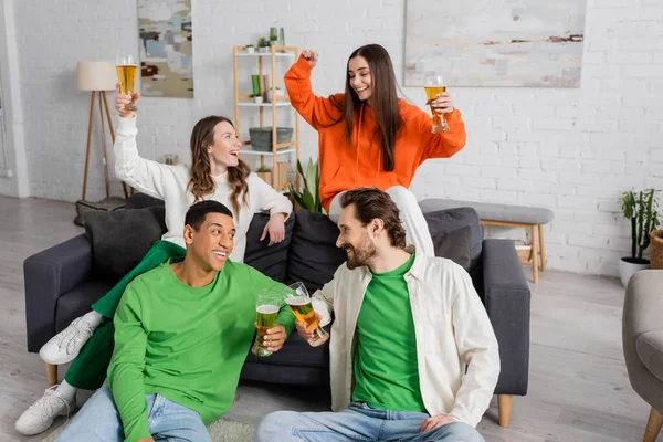 Cheerful multicultural men clinking glasses of beer while sitting near happy women in living room — Foto stock