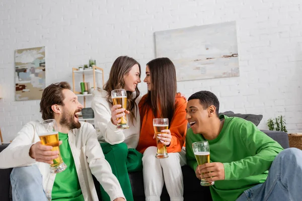Positive and interracial group of friends holding glasses of beer while looking at each other in living room — Stockfoto
