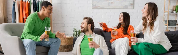 Cheerful woman sitting near friend and pointing at interracial men talking in living room, banner — Stock Photo