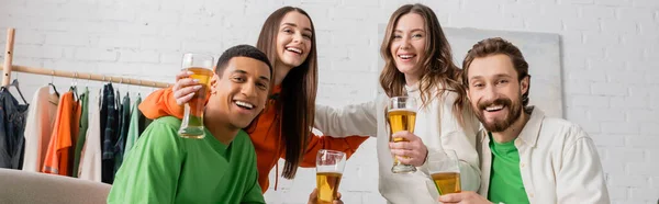 Cheerful women hugging with interracial men holding glasses of beer in living room, banner — Foto stock