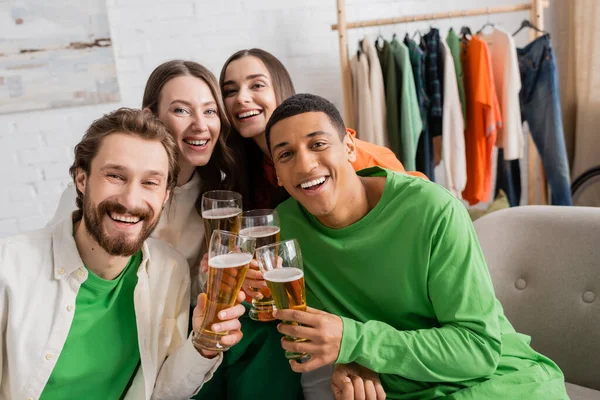 Cheerful women and happy interracial men holding glasses of beer in living room — Stock Photo