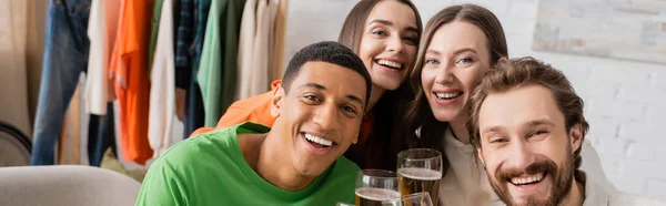 Cheerful women and happy interracial men holding glasses of beer in living room, banner — Stock Photo