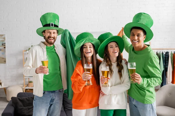 Cheerful and multicultural group of friends in green hats holding glasses of beer while celebrating Saint Patrick Day — Stock Photo