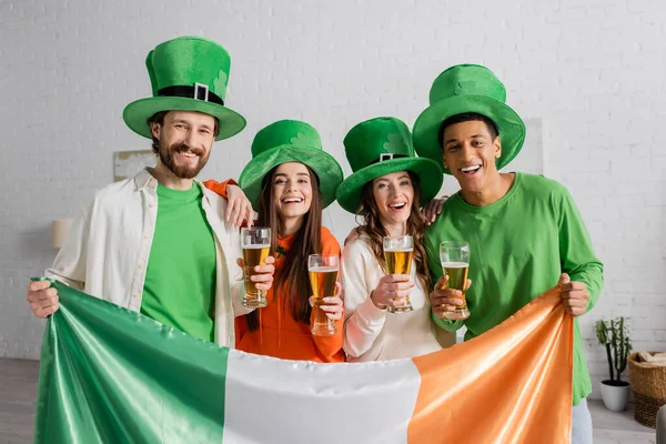 Cheerful and multicultural friends in green hats holding glasses of beer and Irish flag while celebrating Saint Patrick Day — Stockfoto