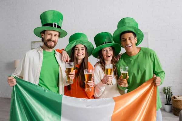 Amigos alegres e multiculturais em chapéus verdes segurando copos de cerveja e bandeira irlandesa enquanto celebra o Dia de São Patrício — Fotografia de Stock