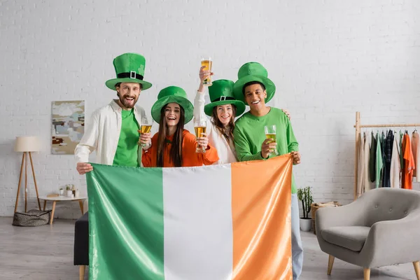 Happy and multicultural friends in green hats holding glasses of beer and Irish flag while celebrating Saint Patrick Day — Stock Photo