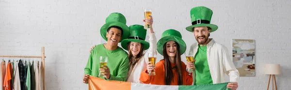 Amici felici e multiculturali in cappelli verdi che tengono bicchieri di birra e bandiera irlandese mentre celebrano il giorno di San Patrizio, banner — Foto stock