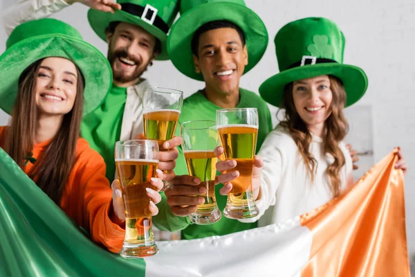 Joyful and multicultural friends in green hats holding glasses of beer and Irish flag while celebrating Saint Patrick Day — Stock Photo