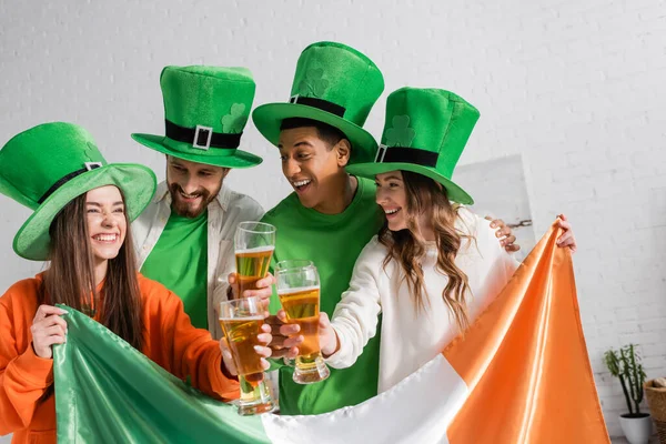 Cheerful and interracial friends in green hats holding glasses of beer and Irish flag while celebrating Saint Patrick Day — Stock Photo