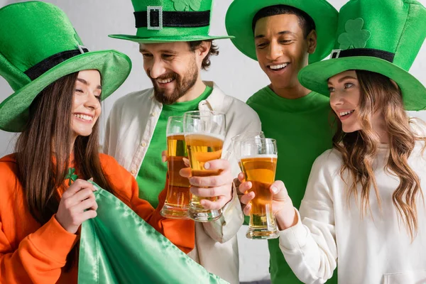 Cheerful and interracial friends in green hats clinking glasses of beer near Irish flag while celebrating Saint Patrick Day — Stock Photo