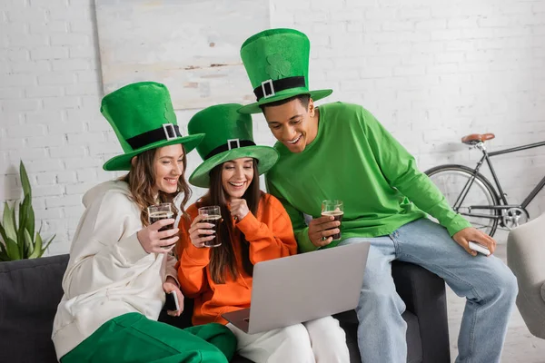 Happy women and african american man in green hats holding glasses of dark beer while looking at laptop — Stock Photo