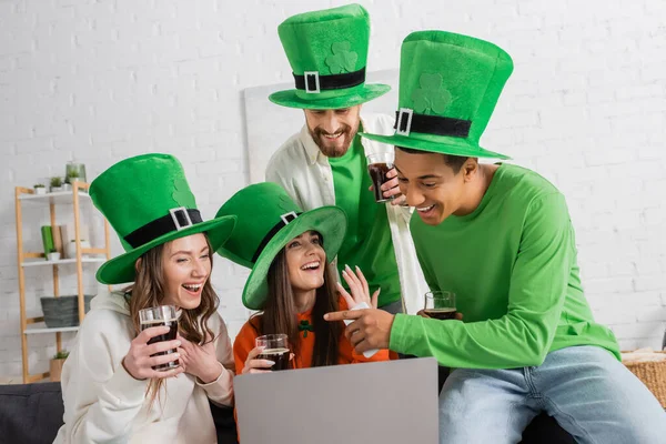 Happy african american man in green hat holding smartphone and pointing at friends during video call on laptop — Stock Photo