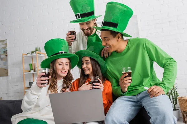 Positive and interracial friends in green hats holding glasses of dark beer while looking at laptop — Stock Photo