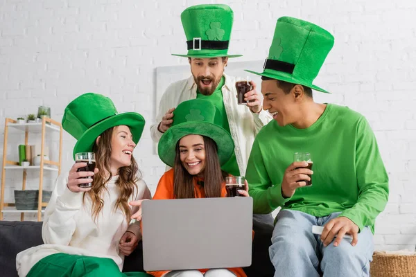 Positive and multiethnic friends in green hats holding glasses of dark beer while looking at laptop — Stock Photo