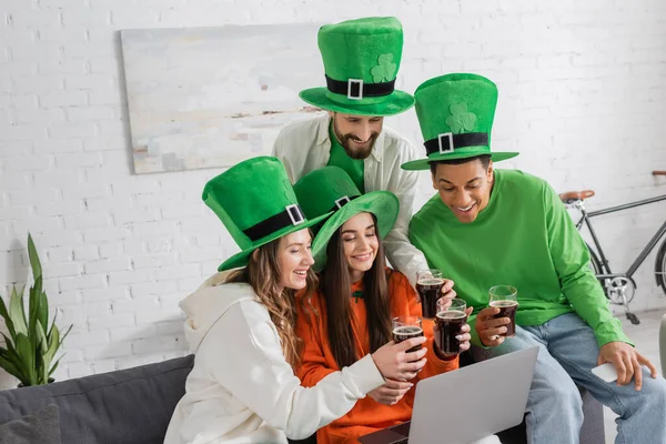 Happy and multiethnic friends in green hats clinking glasses of dark beer while looking at laptop — Stock Photo