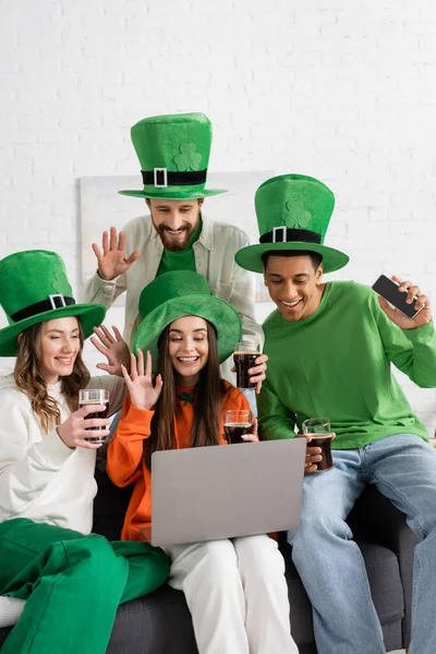 Happy and multiethnic friends in green hats holding glasses of dark beer while waving hands at laptop during video call — Stock Photo