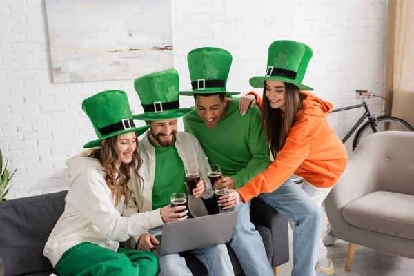 Happy and multiethnic friends in green hats holding glasses of dark beer during video call on laptop on Saint Patrick Day — Stock Photo