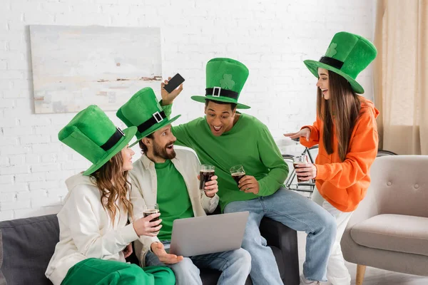 Joyful and multiethnic friends in green hats holding glasses of dark beer during Saint Patrick Day celebration at home — Stockfoto