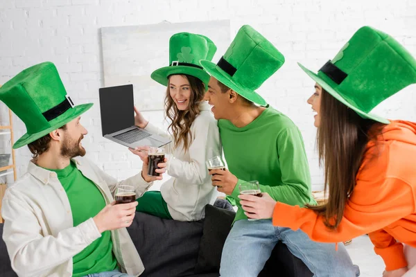 Cheerful woman holding laptop with blank screen near multiethnic friends with glasses of dark beer on Saint Patrick Day — Photo de stock