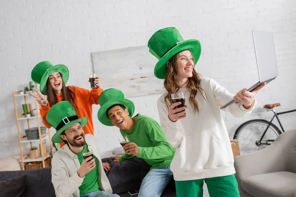 Cheerful woman holding laptop near happy multiethnic friends with glasses of dark beer while celebrating Saint Patrick Day — Stock Photo