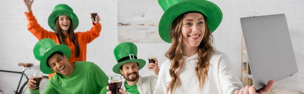 Smiling woman holding laptop near happy multiethnic friends with glasses of dark beer while celebrating Saint Patrick Day, banner — Stock Photo