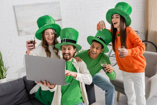 Bearded man holding laptop near joyful multiethnic friends with glasses of dark beer while celebrating Saint Patrick Day — Stock Photo
