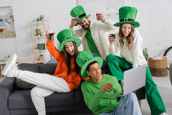 Happy african american man holding laptop during video call near cheerful friends toasting glasses of beer on Saint Patrick Day — Stock Photo