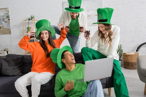 Positive african american man holding laptop during video call near cheerful friends toasting glasses of beer on Saint Patrick Day — Stock Photo