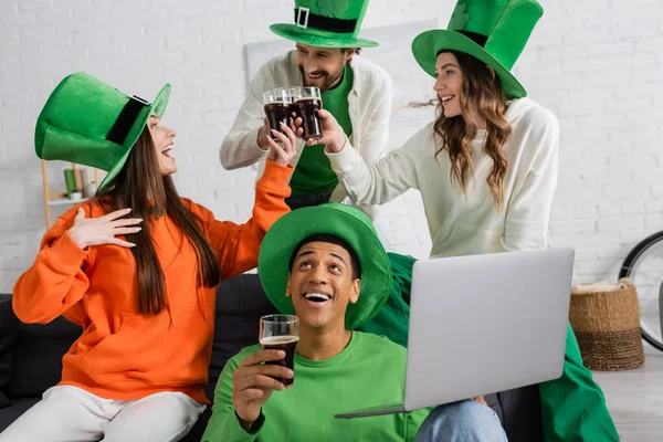 Positive african american man holding laptop while cheerful friends toasting glasses of beer on Saint Patrick Day — Stock Photo