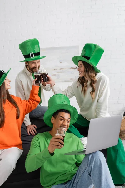 Smiling african american man looking at laptop while cheerful friends toasting glasses of beer on Saint Patrick Day — Stock Photo