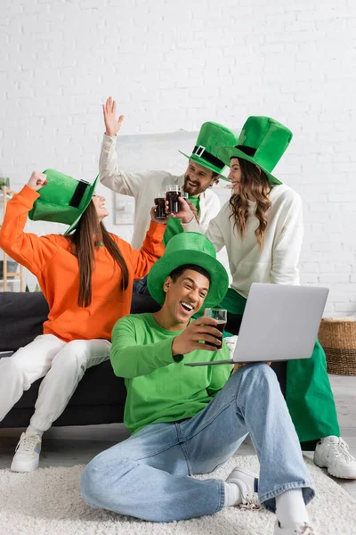Joyful african american man holding laptop during video call near cheerful friends toasting glasses of beer on Saint Patrick Day — Stock Photo