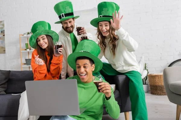 Joyful african american man holding laptop during video call near cheerful friends with glasses of beer on Saint Patrick Day — Stock Photo
