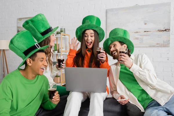 Excitée femme agitant la main lors d'un appel vidéo près d'amis interraciaux dans des chapeaux verts le jour de la Saint Patrick — Photo de stock