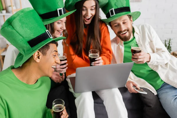 Схвильовані міжрасові друзі дивляться на ноутбук під час відеодзвінка, тримаючи склянки темного пива на День Святого Патрика. — стокове фото