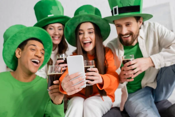 Happy interracial friends looking at smartphone during video call while holding glasses of dark beer on Saint Patrick Day — Stock Photo