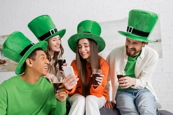 Happy interracial friends looking at friend with smartphone while holding glasses of dark beer on Saint Patrick Day — Stock Photo