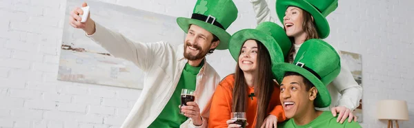 Cheerful bearded man taking selfie with interracial friends holding glasses of dark beer on Saint Patrick Day, banner — Stock Photo